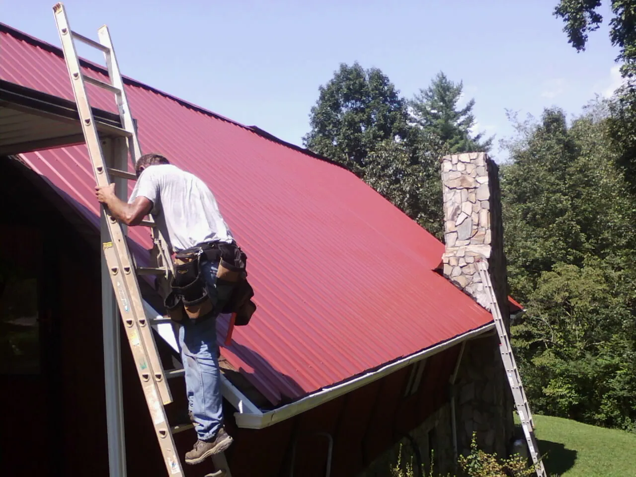 Roofer on a ladder beside a red metal roof and stone chimney — Soffit & Fascia Repair by Roofing Brothers in Inverness